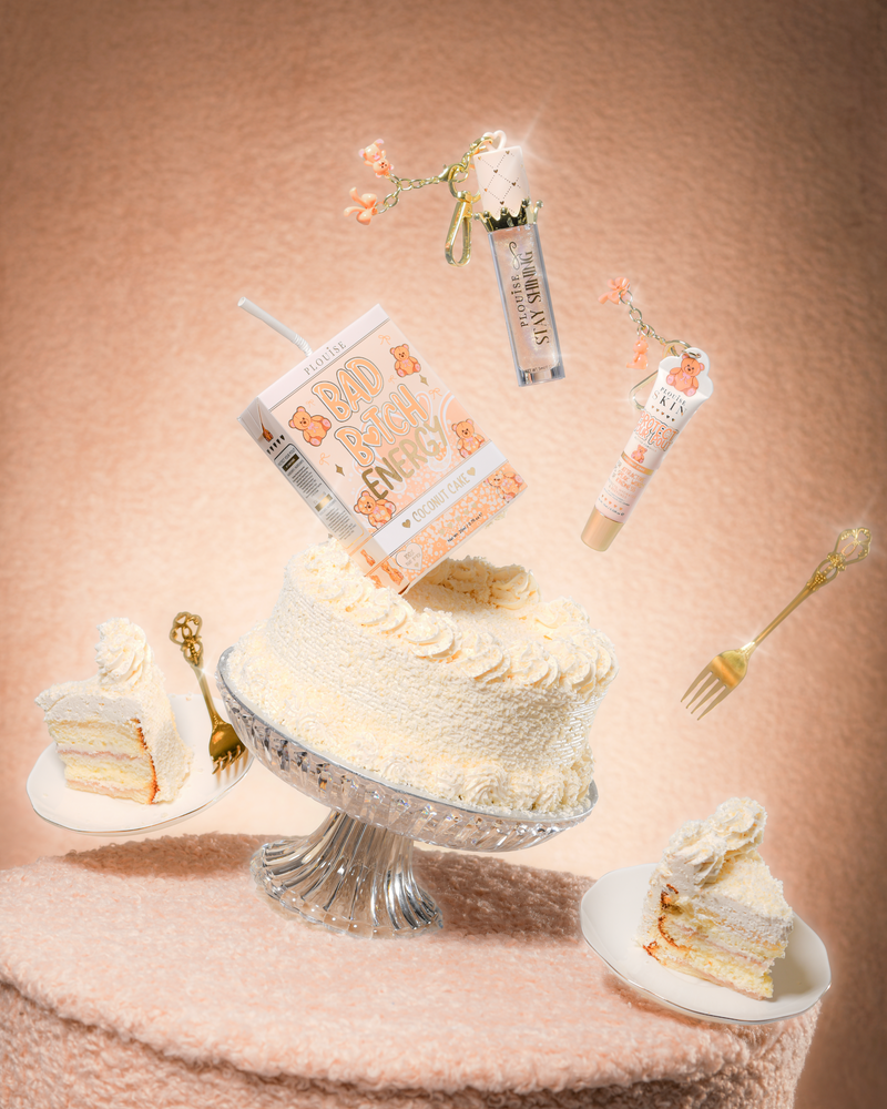 Um bolo em uma mesa de tecido de pelúcia com um P.Louise Teddy Glitter Bad Bitch Energy Lip Duo em bolo de coco contendo um condicionador labial e óleo labial flutuando ao redor do bolo
