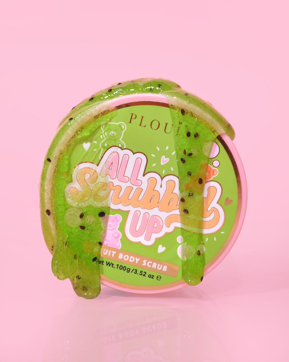 Um recipiente de P.Louise All Scrubbed Up Body Scrub com o rótulo 'Candy Bears' em um fundo rosa, com o produto aparecendo como um gel verde