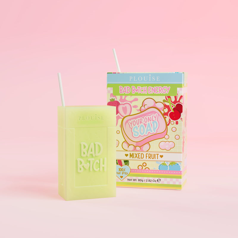 Uma barra de sabonete verde com 'Bad Bitch' gravada, acompanhada por uma caixa de suco colorida com a inscrição 'Mixed Fruit', apresentada em fundo rosa.