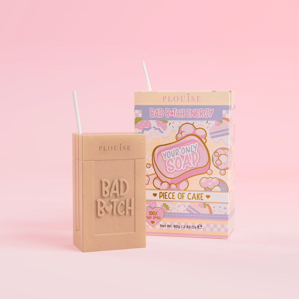 Una pastilla de jabón beige con la palabra "Bad Bitch" grabada, acompañada de una colorida caja de jugo con la etiqueta "Piece of Cake", presentada sobre un fondo rosa.