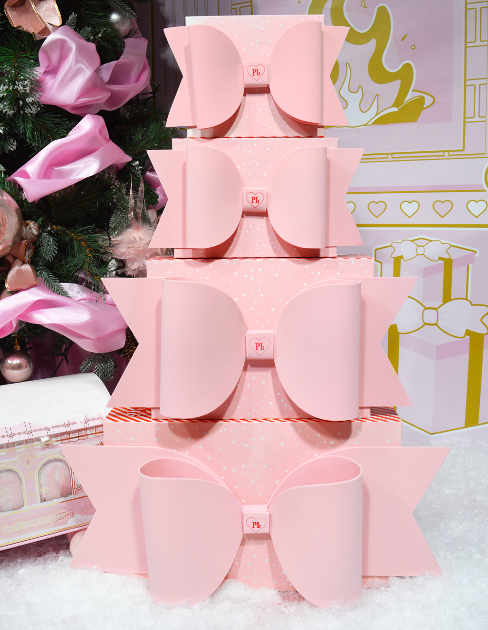 Pilha de caixas de embrulho rosa P.Louise com laços exibidas em um cenário de Natal com decorações.
