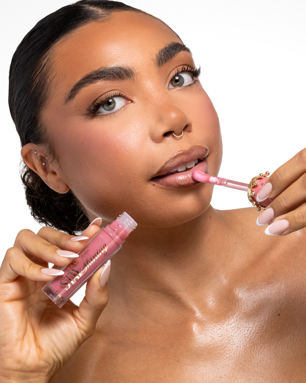 Un primer plano de una mujer aplicándose un producto para labios líquido P.Louise Stay Shining Lip Oil rosa con un aplicador de pie mientras sostiene el tubo en el otro. mano, sobre un fondo blanco limpio.