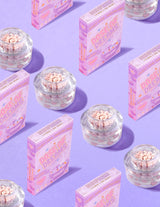 Envases transparentes de P.Louise DOse of Dreams Viatmin C Capsule Face Cream con una cápsula rosa rodeada de cajas de cereales rosas sobre un fondo morado