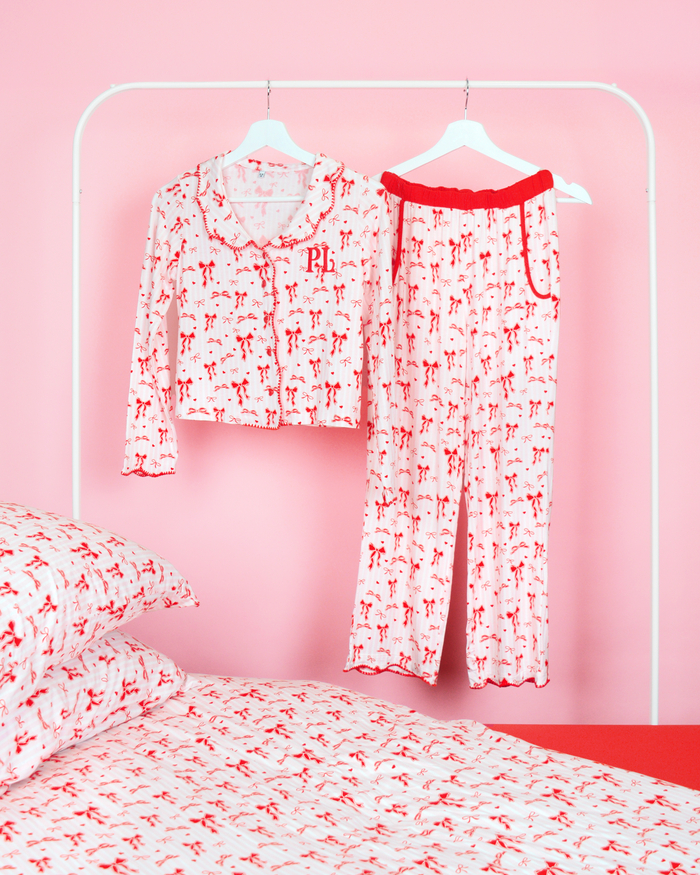 Conjunto de calças de pijama com laço de Natal para meninas P.Louise com um desenho de laço vermelho contra listras rosa e brancas penduradas em cabides em um quarto rosa com roupa de cama combinando