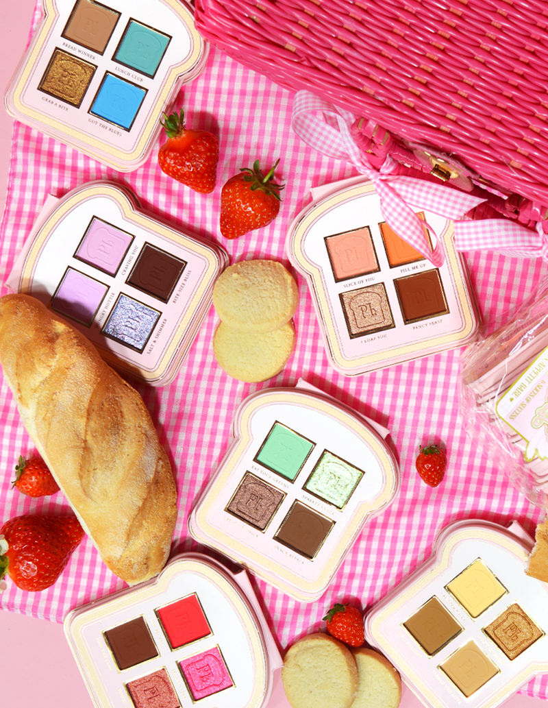 Um conjunto de mini paletas de sombras estilo sanduíche com vários tons de maquiagem exibidos em uma superfície de algodão rosa, acompanhadas de morangos e um pedaço de pão.