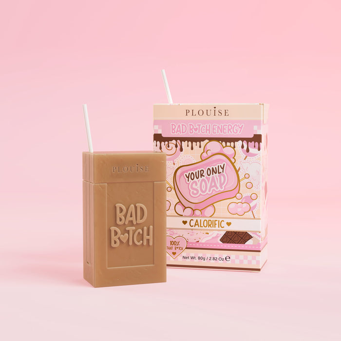 Uma barra de sabonete marrom com 'Bad Bitch' gravado, acompanhada por uma caixa de suco colorida com o rótulo 'Calorific', apresentada em fundo rosa.