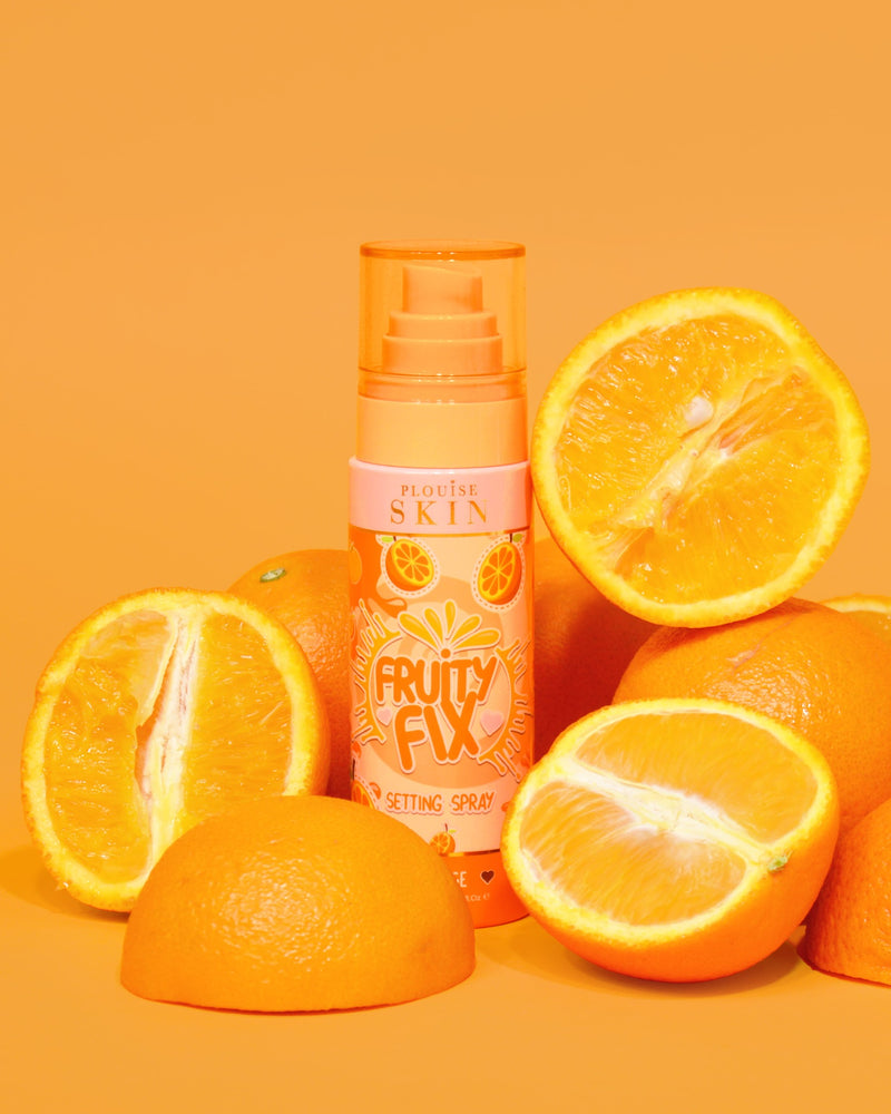 Um frasco laranja de P. Louise Fruity Fix Vitamin C Setting Spray com um fundo laranja rodeado por fatias de laranja fresca.