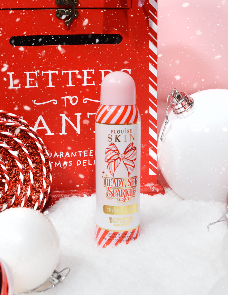 Uma garrafa de P. Louise Ready Set Sparkle Christmas Glitter Setting Spray cercado por decorações festivas como neve, bugigangas e uma caixa de correio de Natal