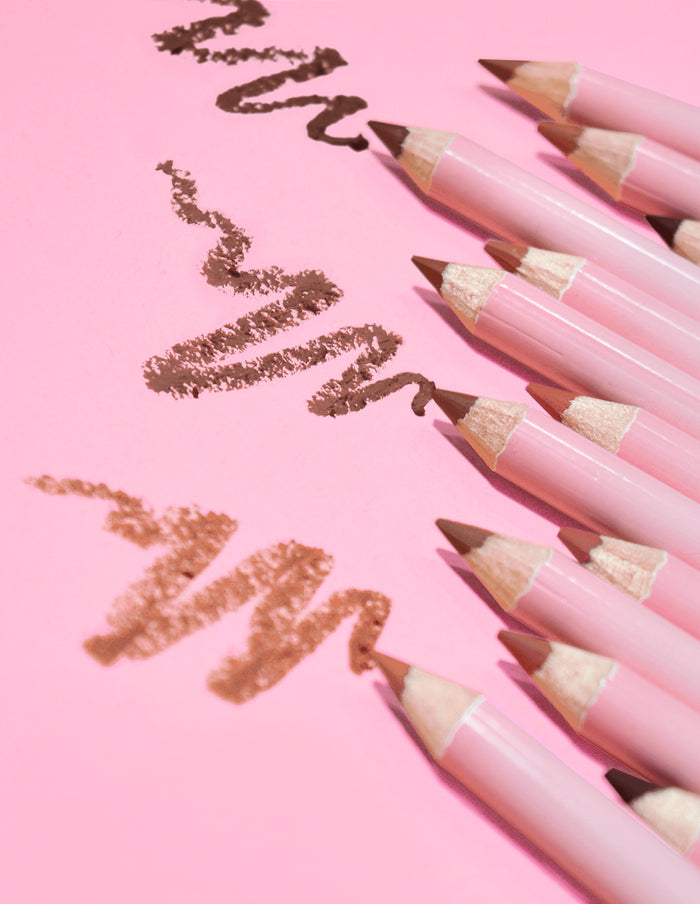 Uma coleção de delineadores labiais de última definição P.Louise em uma variedade de tons nude, dispostos sobre um fundo rosa com algumas linhas do produto desenhadas na superfície.
