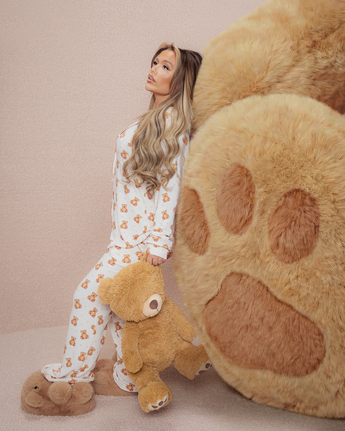 Mujer en pijama de peluche sosteniendo un osito de peluche junto a una gran pata de oso de peluche sobre un fondo beige