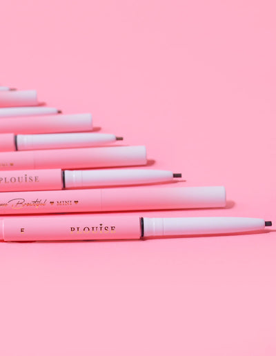Uma coleção de Mini Lápis de Sobrancelha P.Louise dispostos em uma fileira com o nome da marca visível no corpo dos lápis, exibidos contra um fundo rosa